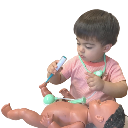A child in a pink shirt uses a toy thermometer on a doll's foot while wearing a toy stethoscope around their neck.