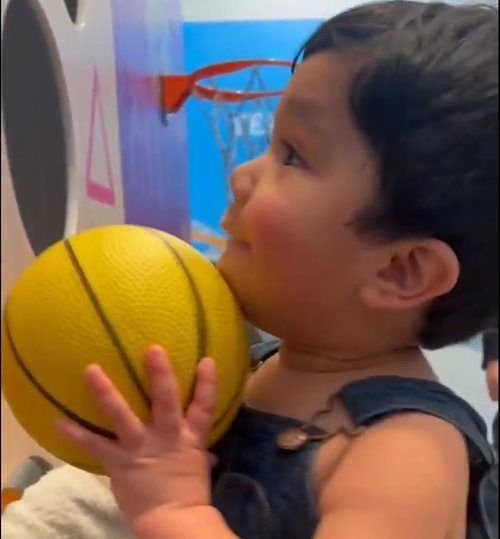 A young child holding a yellow basketball near a toy hoop, looking up with a smile.