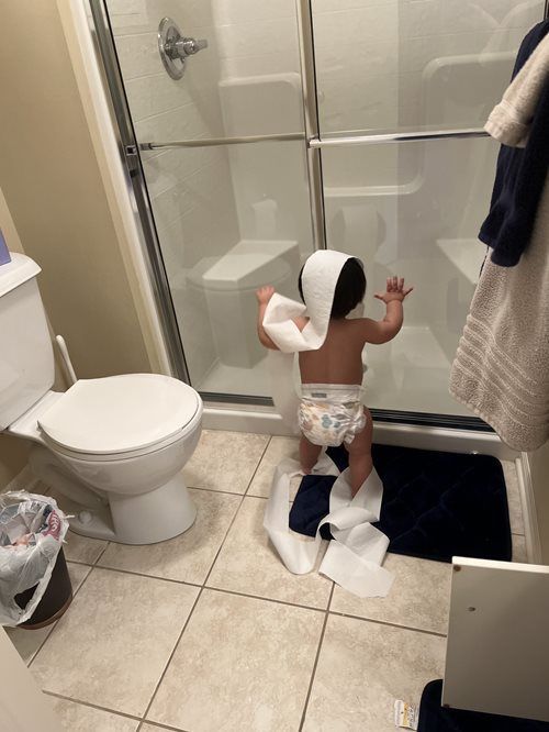 A small child in a diaper stands in a bathroom, draped in toilet paper while reaching toward the shower glass door.