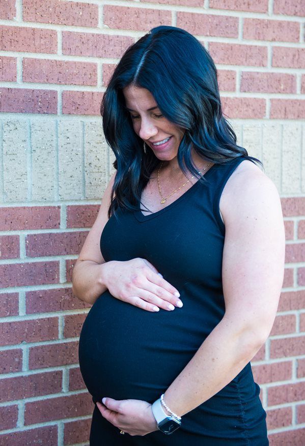 A person in a black tank top stands before a brick wall, looking down and cradling their pregnant belly with both hands.