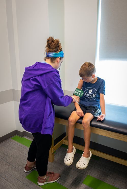 A healthcare professional in a purple hoodie places a blood pressure cuff on a patient's arm in a clinical exam room.
