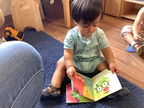 A child in a blue outfit sits on a blue rug, looking down while reading an illustrated board book with monster characters.