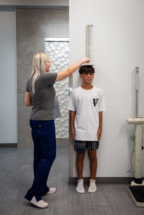 A healthcare worker uses a wall-mounted stadiometer to measure the height of a patient standing against a white wall.