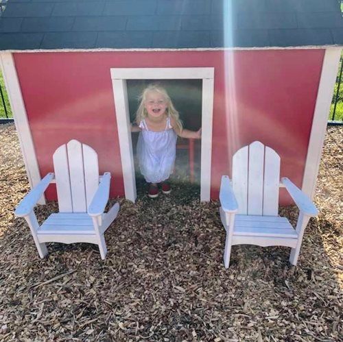 A smiling child in a light-colored dress stands inside a small, red playhouse with two white chairs in the foreground.