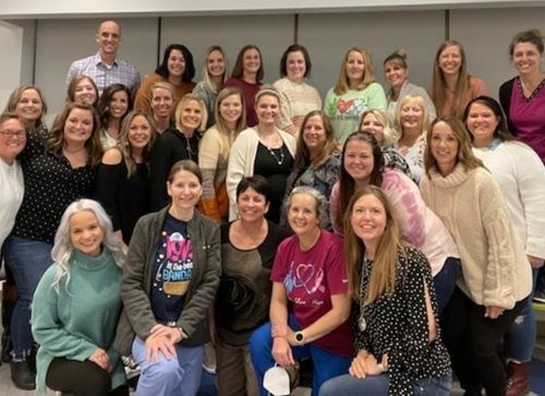 A large group of people smiling for a photo indoors, posed in rows against a gray wall.