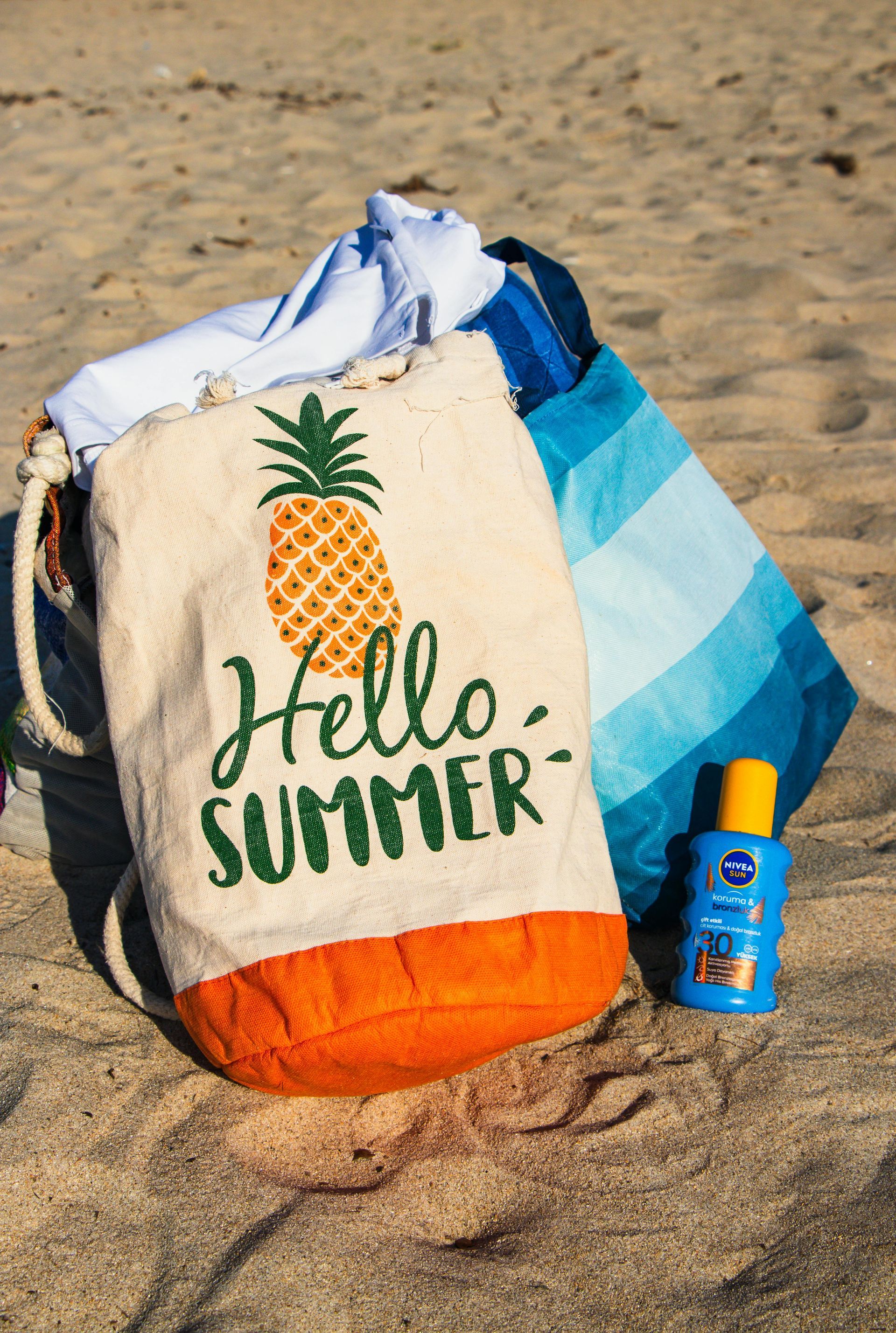 Sandy area with a bag with a picture of a pineapple and hello summer on it sitting next to sunscreen