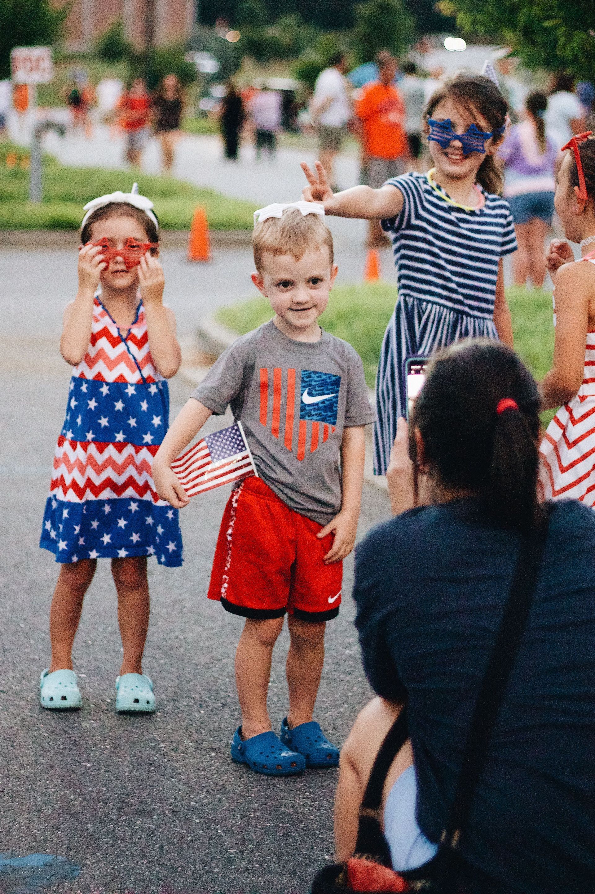 Children waving at a street parade, with an adult kneeling in front of them taking a picture.