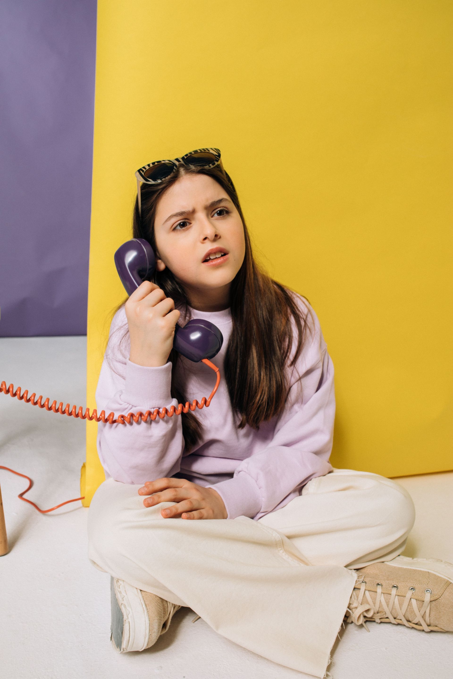 Girl sitting against a yellow and purple backdrop, holding a purple corded phone and looking concerned.