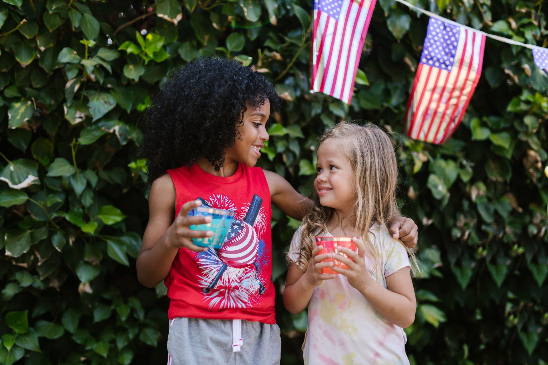 Two children holding drinks and smiling outdoors with American flags and leafy greenery behind them