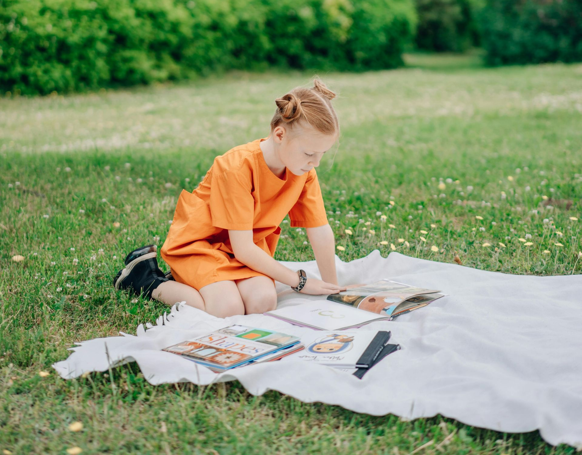 Girl in orange outfit reading on a white blanket in a grassy park