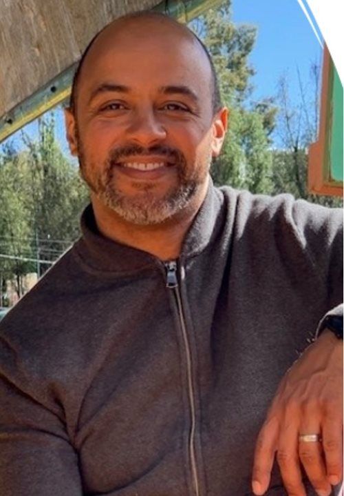 A smiling person with a shaved head and a light beard, wearing a dark gray zip-up jacket outdoors in a sunny setting.