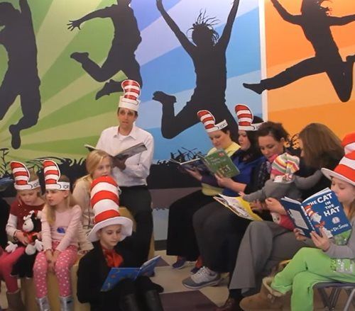A group of children and adults wearing Cat in the Hat striped hats, reading books in a room with colorful wall art.