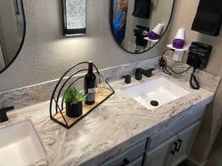 A bathroom with two sinks , two mirrors and a bottle of wine on the counter.