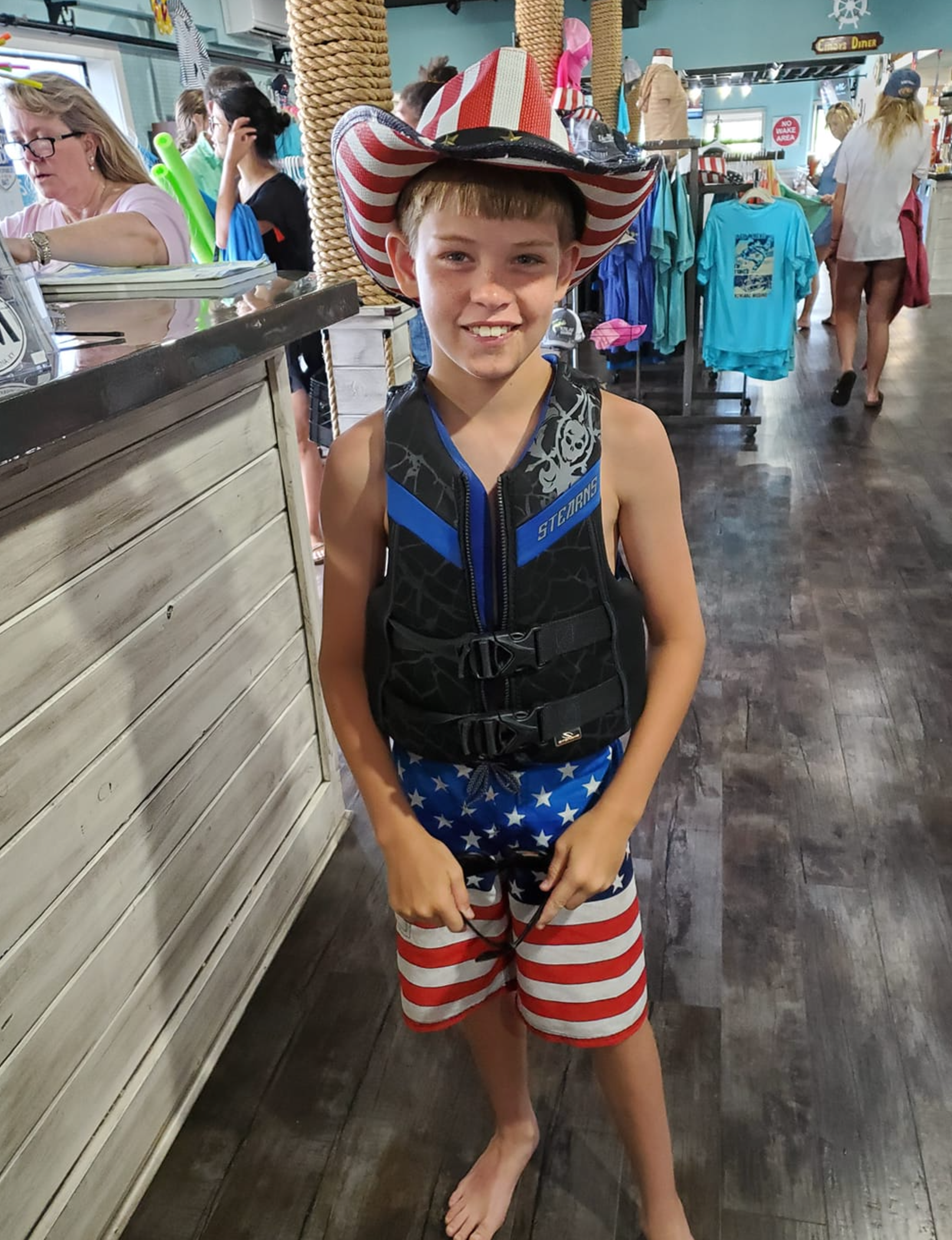 A young boy wearing a life jacket and a cowboy hat is standing in a store.