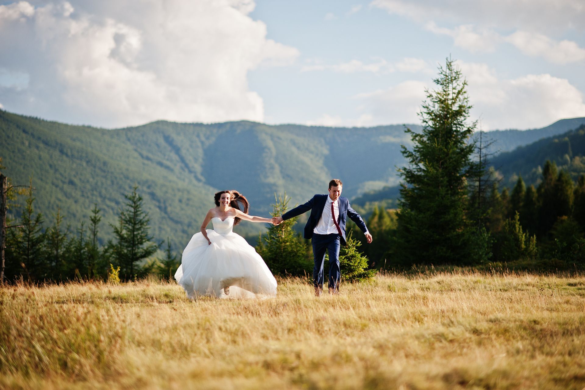 A bride and groom are running through a field holding hands.
