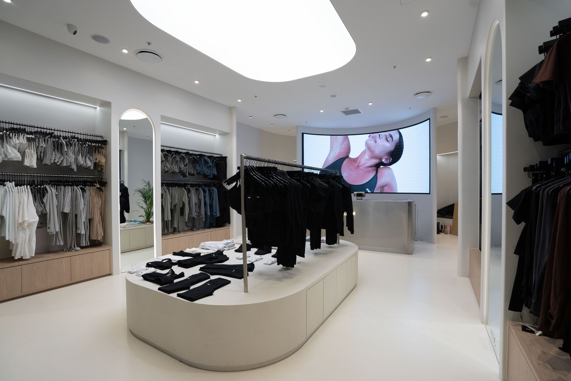 Interior view of a modern clothing store with a large screen displaying a woman; clothes are displayed on racks and shelves.