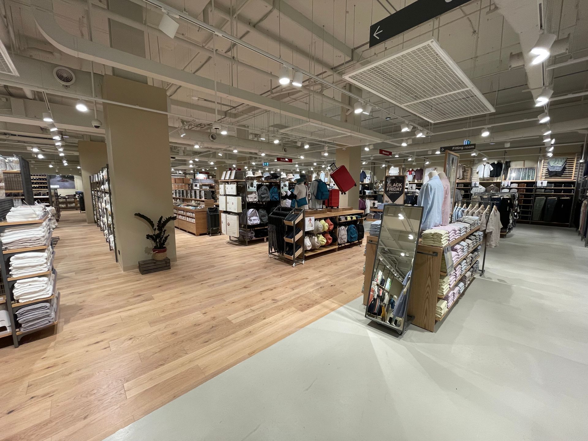 Inside a well-lit store, customers browse merchandise displayed on shelves and tables. Wooden floors and neutral-colored pillars.