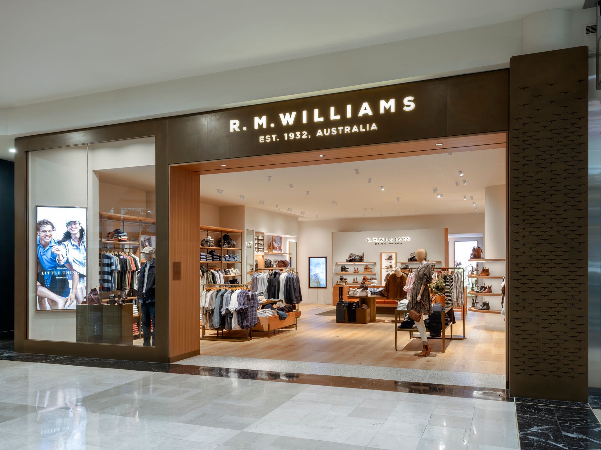 R.M. Williams store entrance. Brown exterior with illuminated logo. Inside displays clothing and accessories on wooden fixtures.