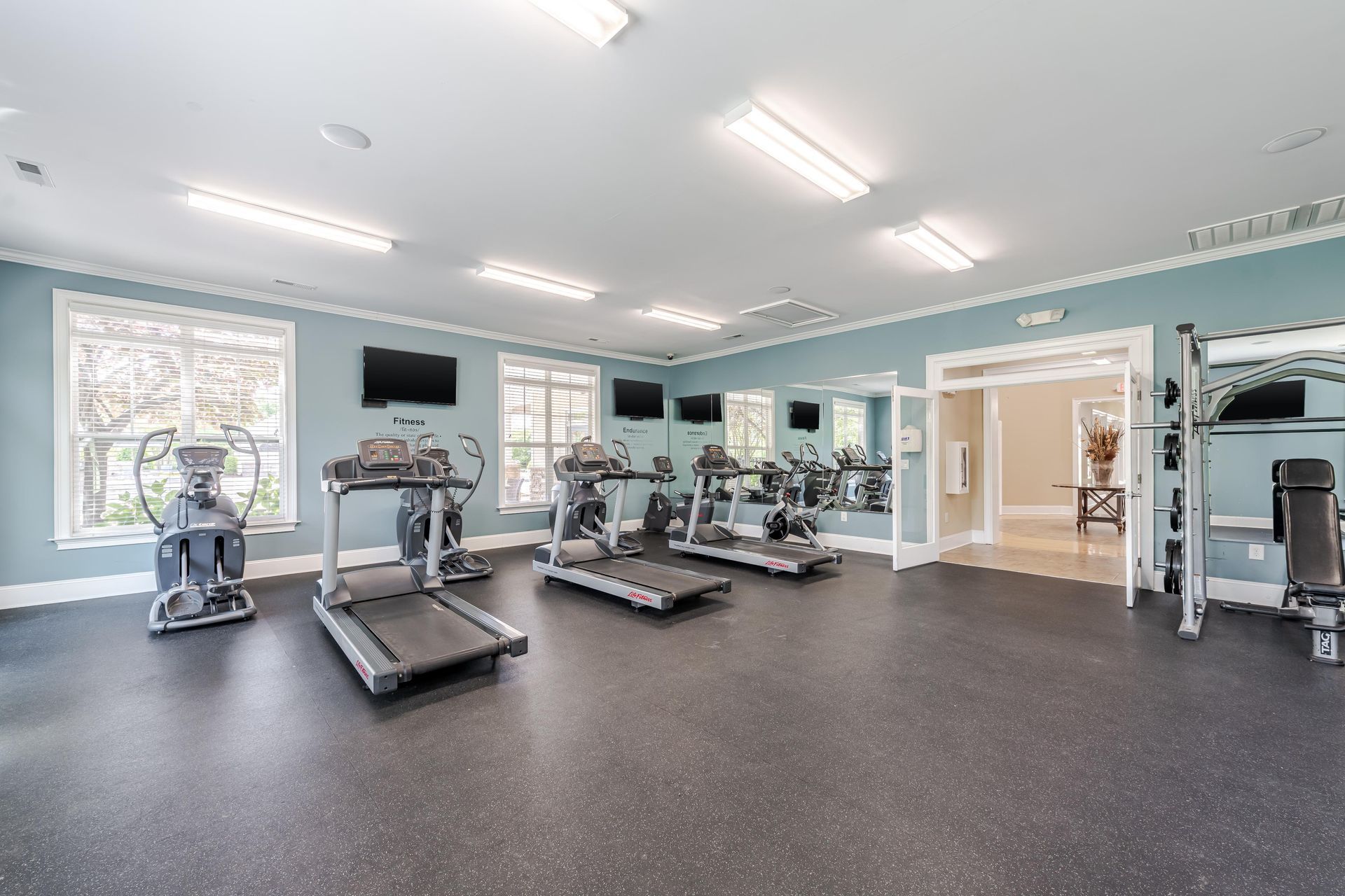 Gym with treadmills, elliptical, exercise equipment, dark floor, light blue walls, and windows at Plantation at Pleasant Ridge in Greensboro, NC.