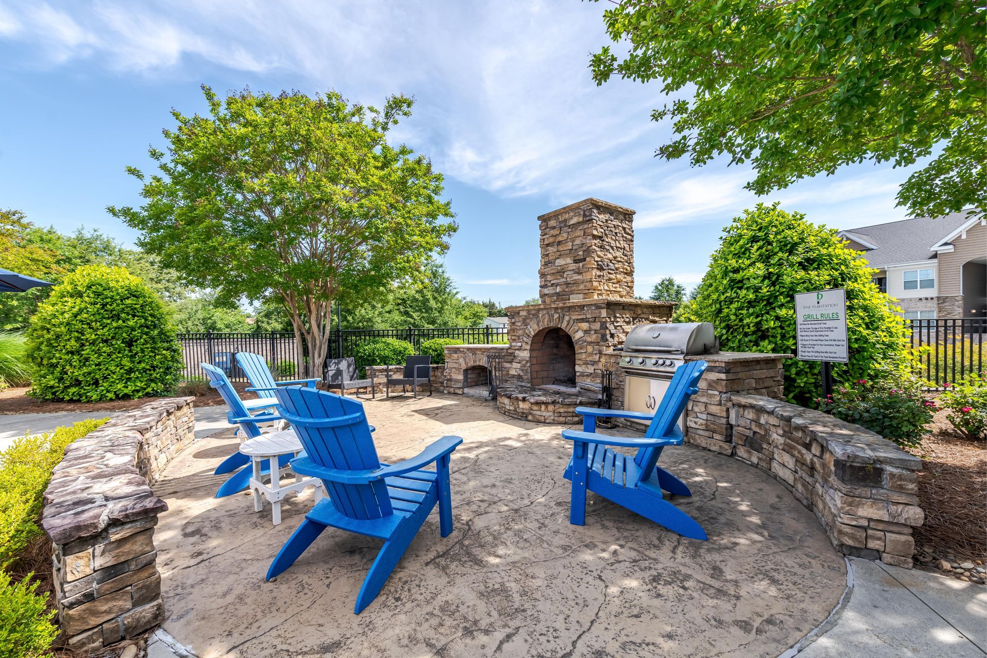 Outdoor patio with blue chairs, stone fireplace, and grill, sunny day at Plantation at Pleasant Ridge in Greensboro, NC.