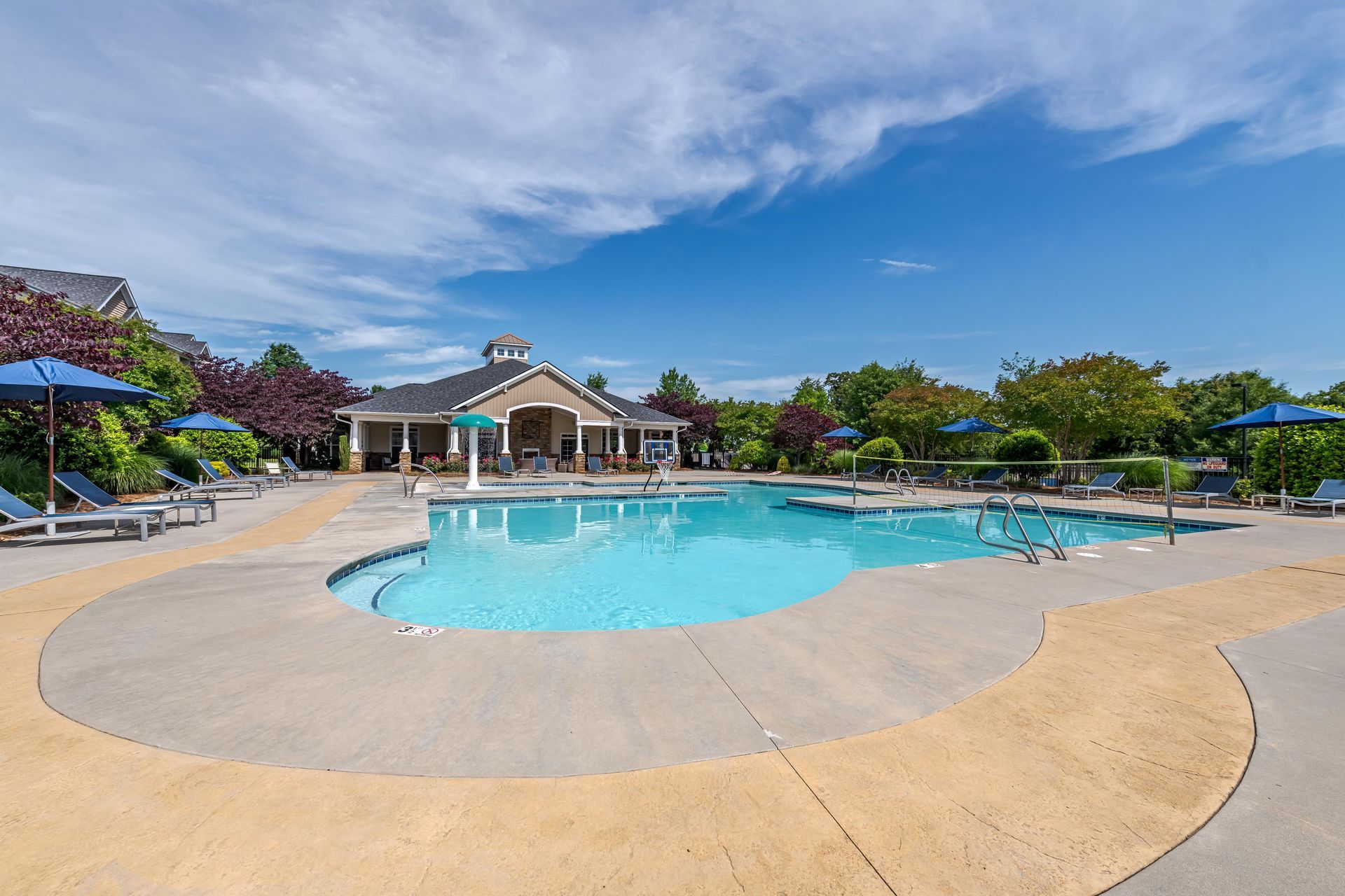 Swimming pool surrounded by concrete deck, lounge chairs, blue umbrellas, and a clubhouse under a blue sky at Plantation at Pleasant Ridge in Greensboro, NC.