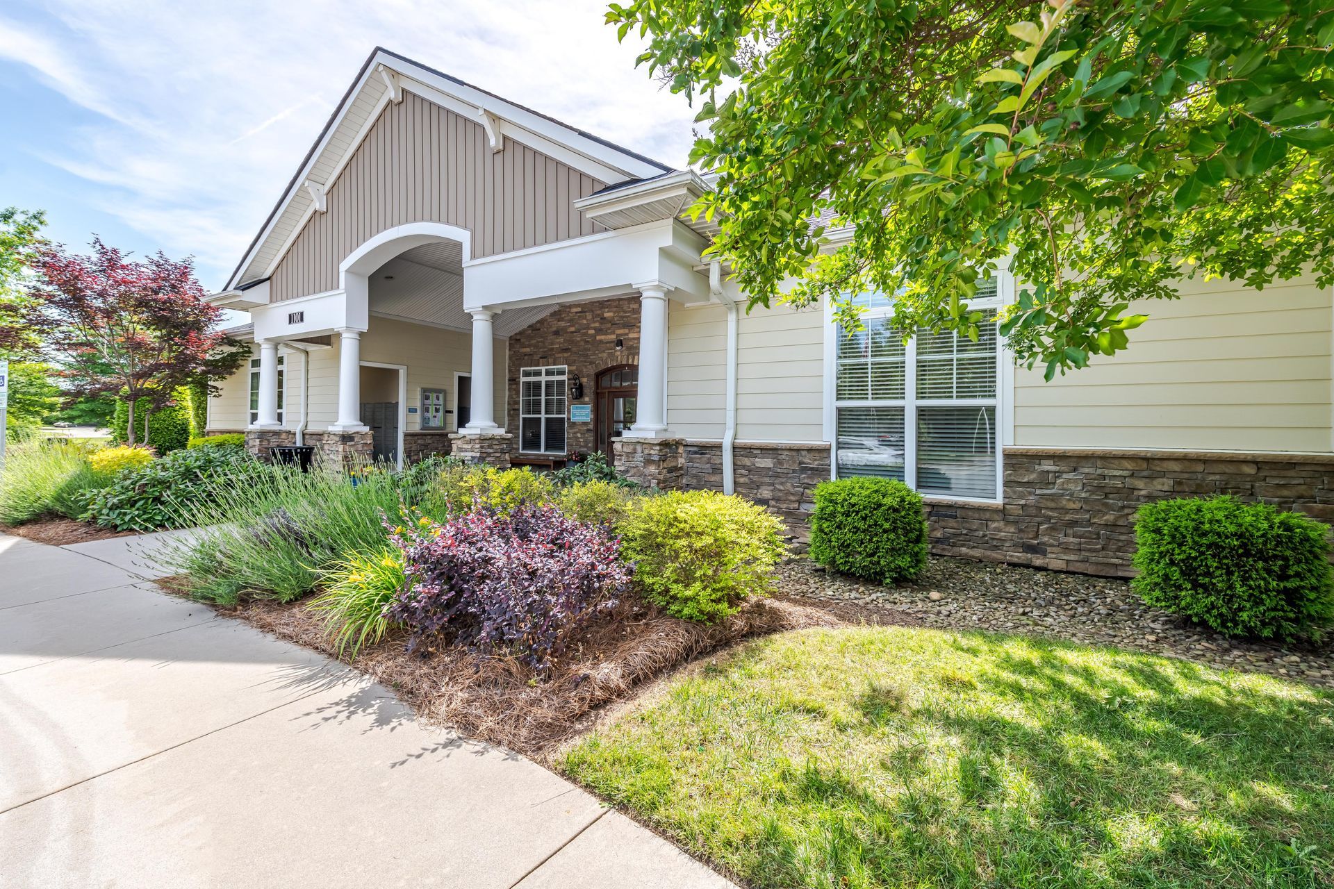 Beige and stone building with a covered entrance, lush landscaping, and green lawn on a sunny day at Plantation at Pleasant Ridge in Greensboro, NC.
