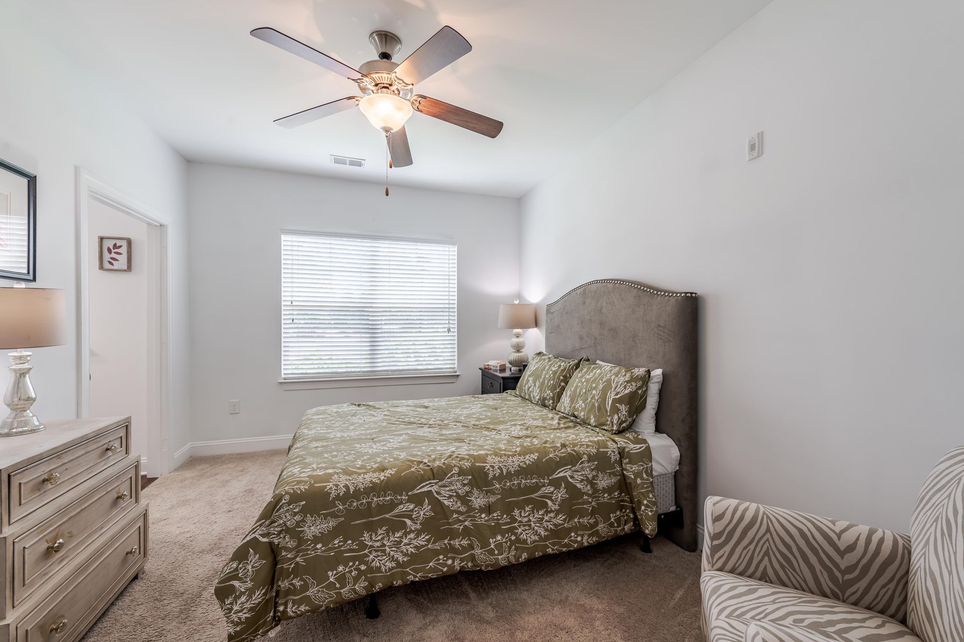 Bedroom with a bed, dresser, chair, and a ceiling fan; neutral tones at Plantation at Pleasant Ridge in Greensboro, NC.