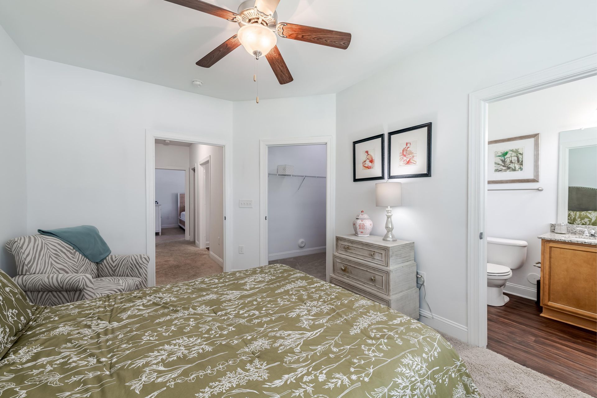Bedroom with a patterned bedspread, a patterned chair, a dresser, and three doorways at Plantation at Pleasant Ridge in Greensboro, NC.