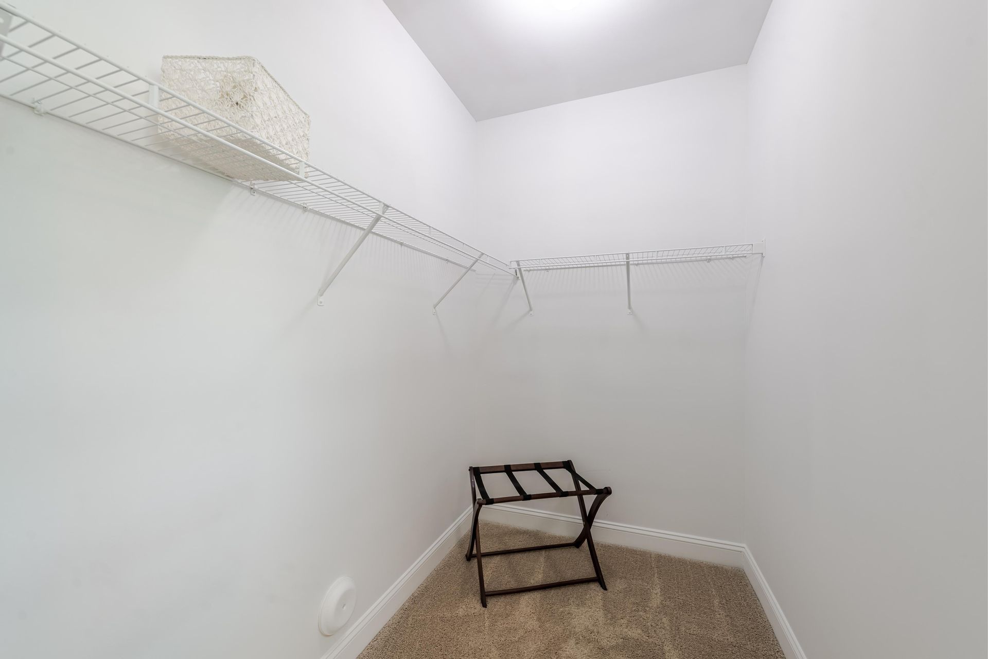 Empty white walk-in closet with wire shelving, beige carpet, and a folding luggage rack at Plantation at Pleasant Ridge in Greensboro, NC.