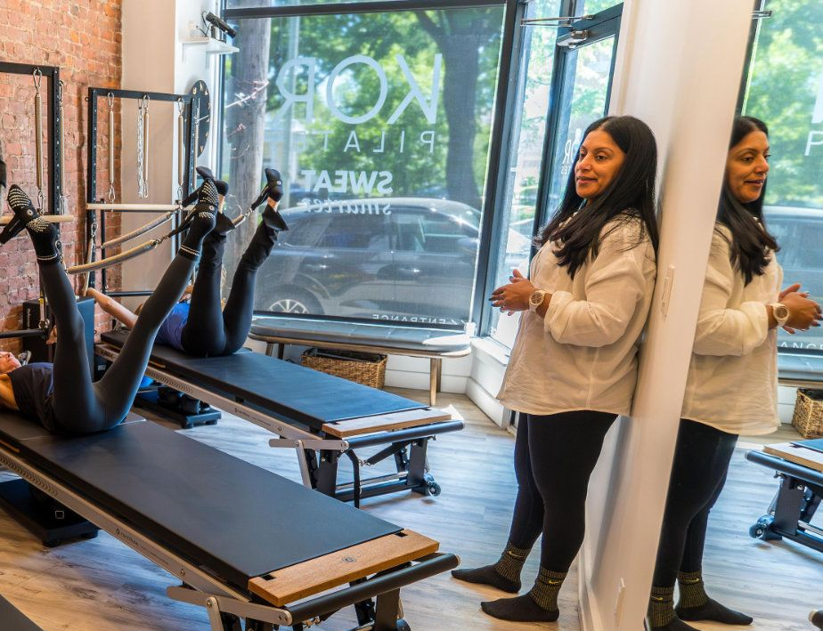 Woman standing by Pilates reformer machines in a bright studio with large windows.