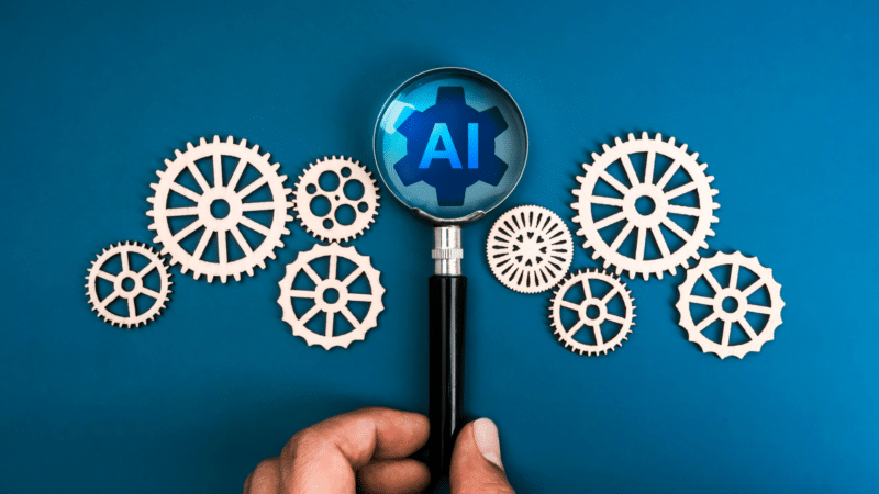 Hand holding magnifying glass over AI icon within gears on blue background.