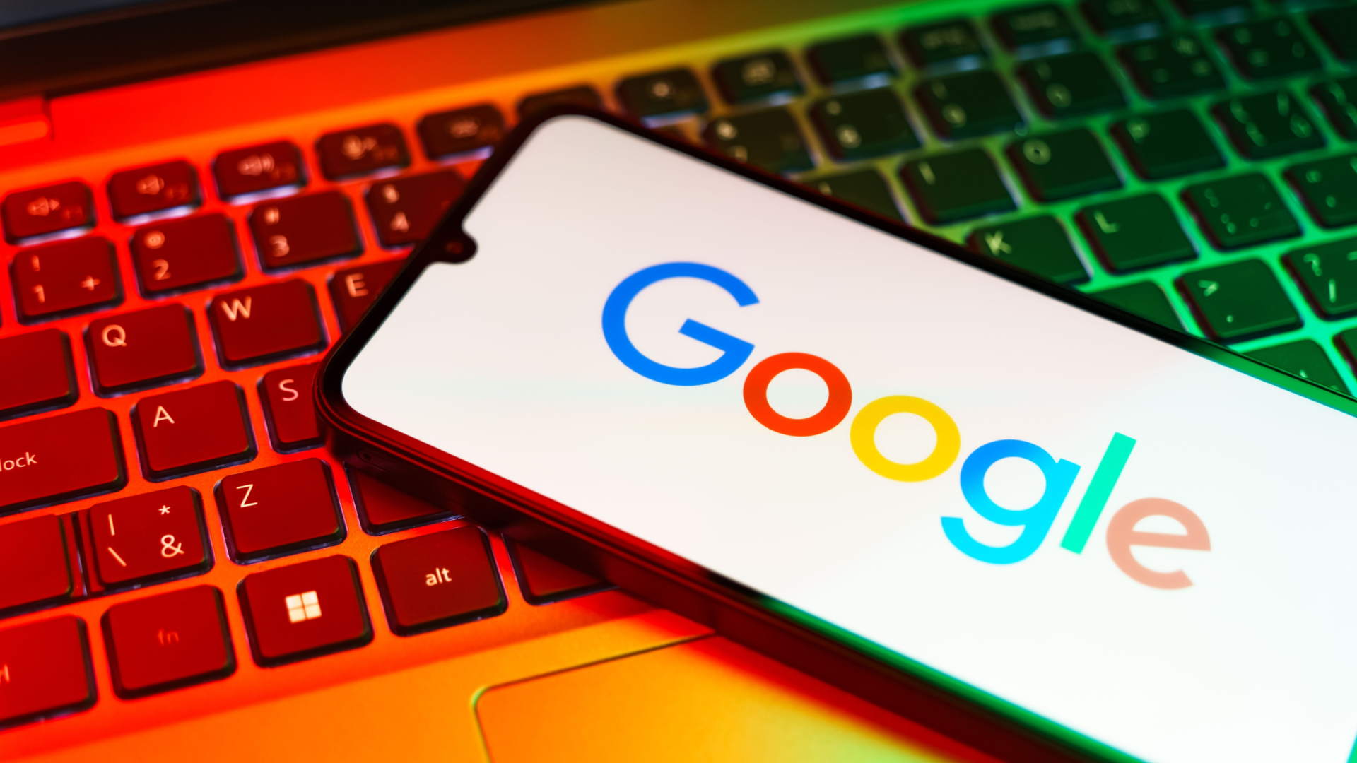 Smartphone displaying the Google logo, resting on a laptop keyboard with colorful lighting.