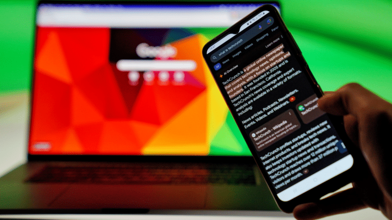 Person holding a phone displaying text with a laptop in the background, both with Google interfaces.