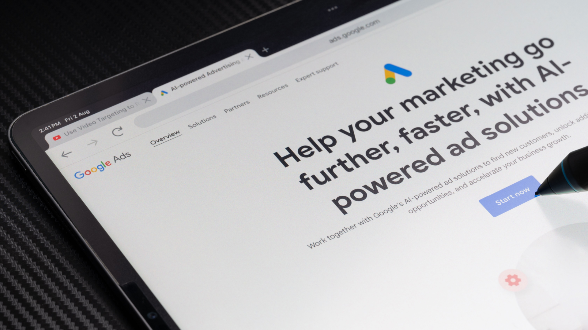 A tablet screen showing the Google Ads webpage, with a hand and stylus interacting with it.