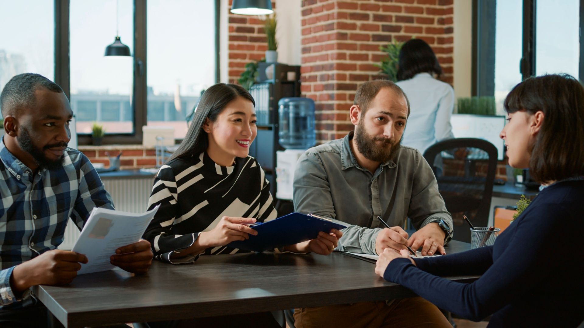 Four diverse colleagues interview a woman at a table in a modern office with brick walls.