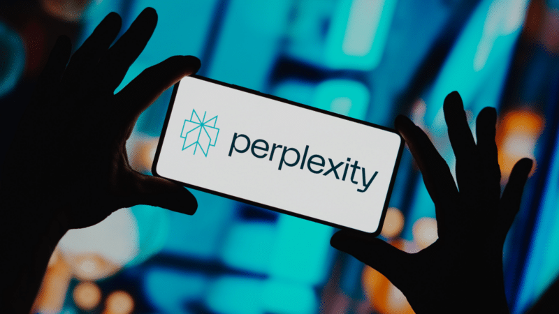 Hands holding a phone displaying the Perplexity AI logo against a blurred, blue-toned backdrop.