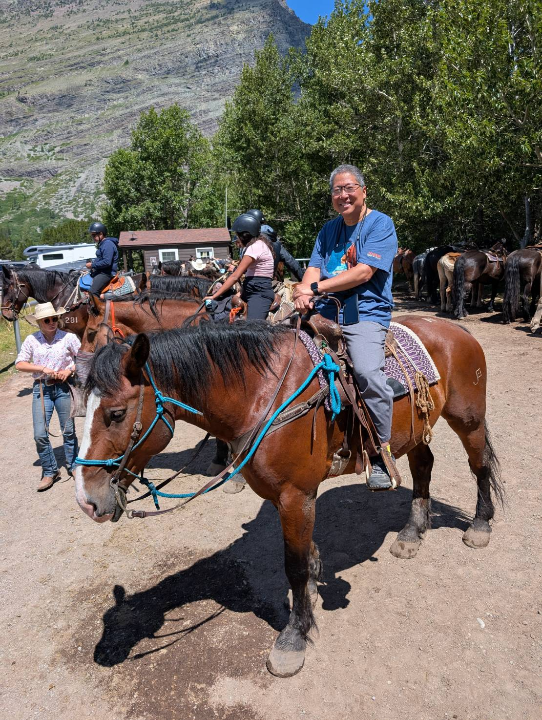 Man on brown horse, smiles at camera. Others ride horses in outdoor setting with mountains.