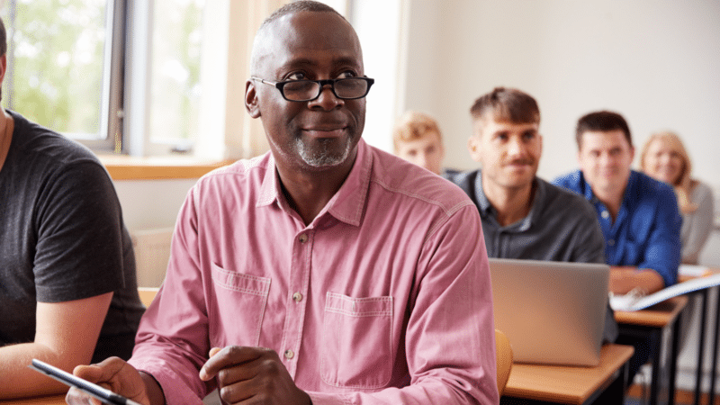 Black man with glasses in a classroom smiles, holding a tablet, seated with other students.