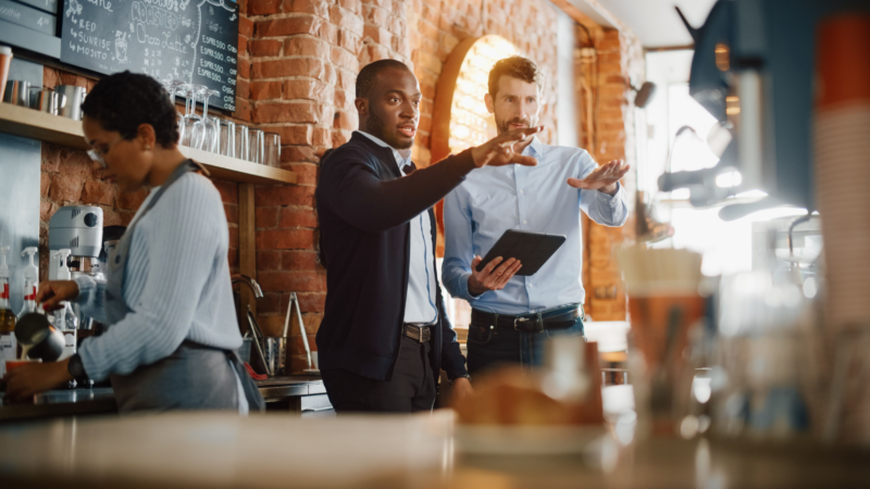 Two men in suits discussing something in a cafe, while a barista works behind the counter.