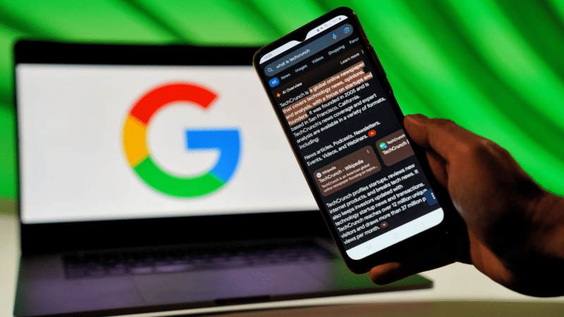 Person holding phone displays news feed, Google logo on laptop in background.