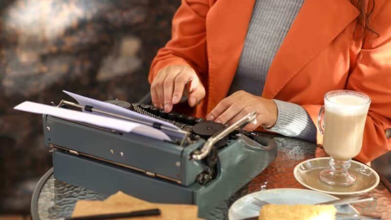 Person typing on a vintage typewriter outdoors, wearing orange jacket, with latte and cake on the table.