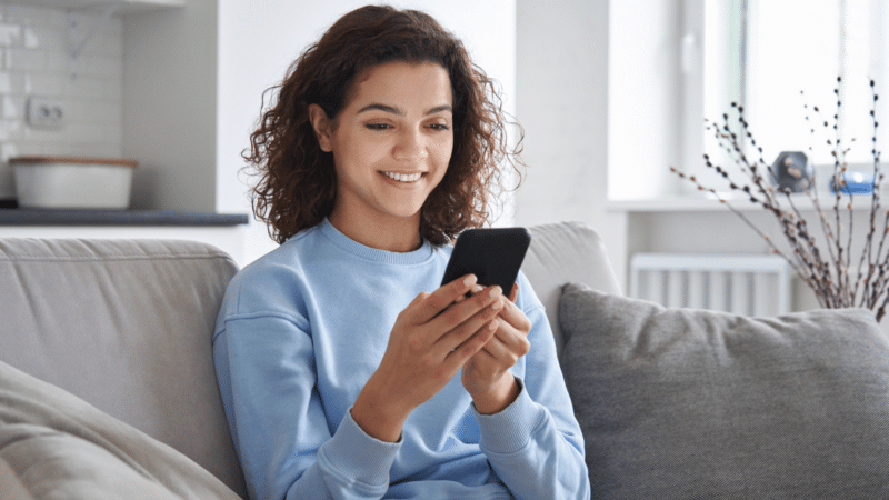 Smiling woman with curly hair using a smartphone while sitting on a couch.