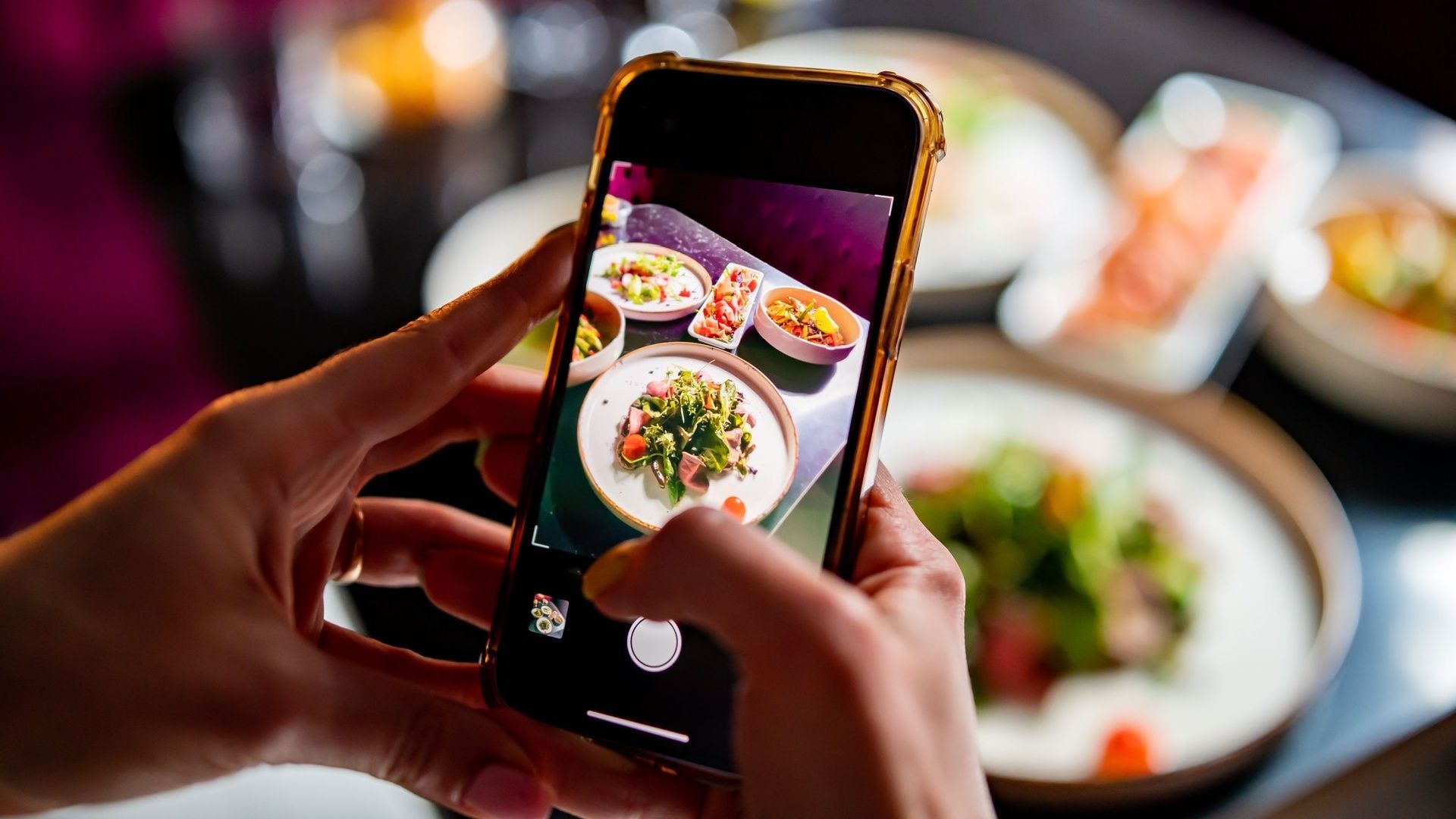 Hands holding a phone, taking a photo of a colorful plate of food at a restaurant table.