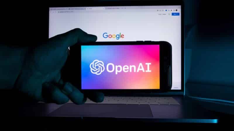 Hand holding a smartphone displaying the OpenAI logo against a laptop screen with the Google search engine.