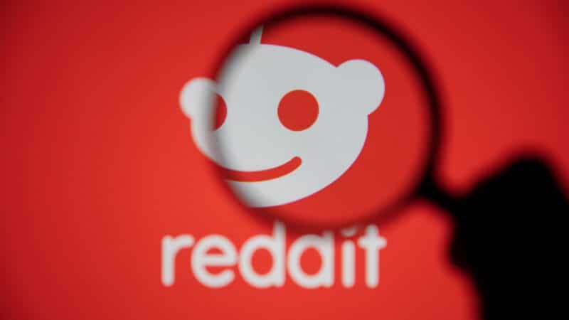Reddit logo, white alien head on red background, magnified by a black-handled magnifying glass.