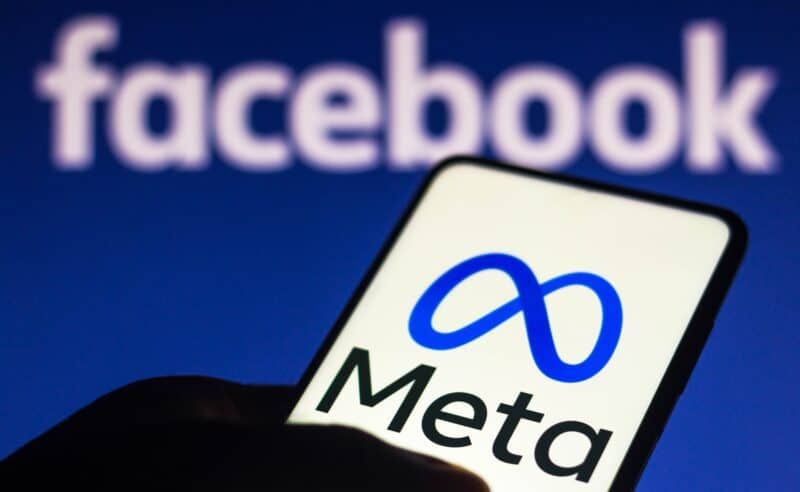 Hand holding a phone with the Meta logo in front of a blurred Facebook logo.