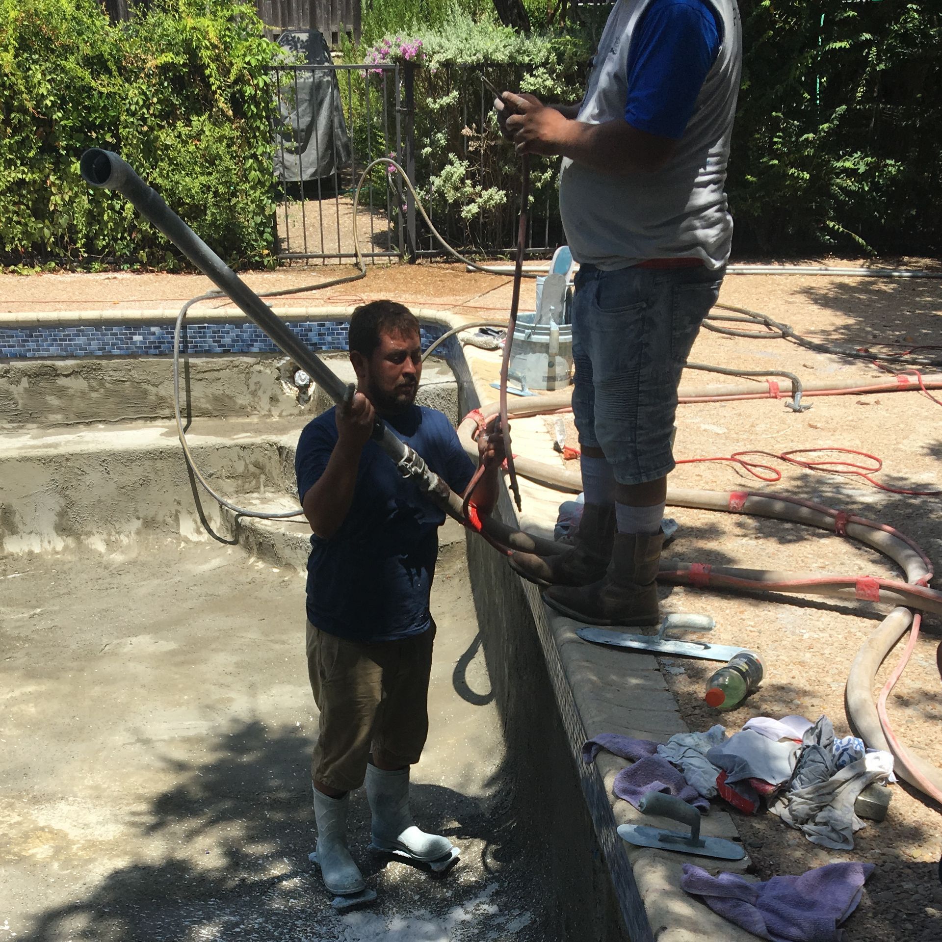 Two men repair a pool, using tools. One holds a pole; the other stands on the pool's edge. Outdoors, sunny.