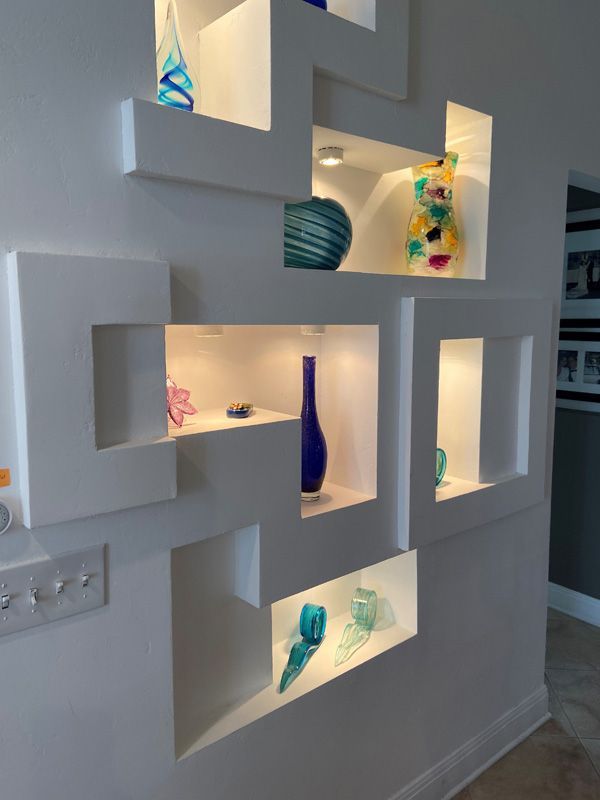 White wall with geometric niches, lit and displaying colorful glass art.