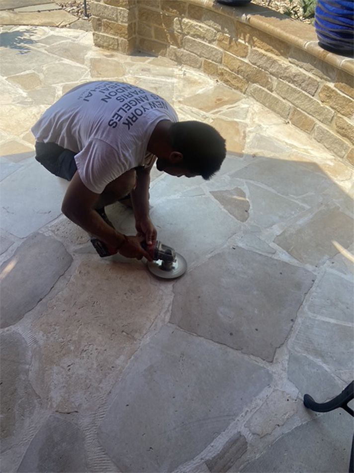 Person kneeling, using a tool to work on stone patio tiles.