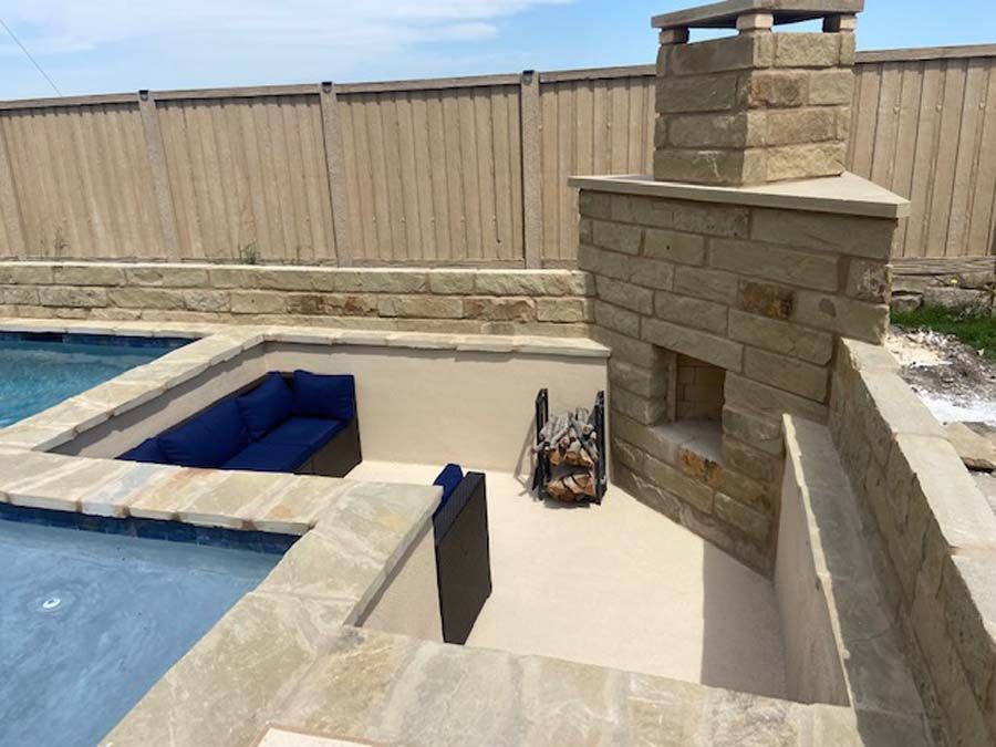 A backyard patio with a pool, seating, and a stone fireplace against a wooden fence.